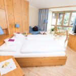 Ein Hotelzimmer mit einem Doppelbett, Holzmöbeln, einem Fernseher, einer kleinen Sitzecke an großen Fenstern und blauen Akzenten in Lampen und Vorhängen.