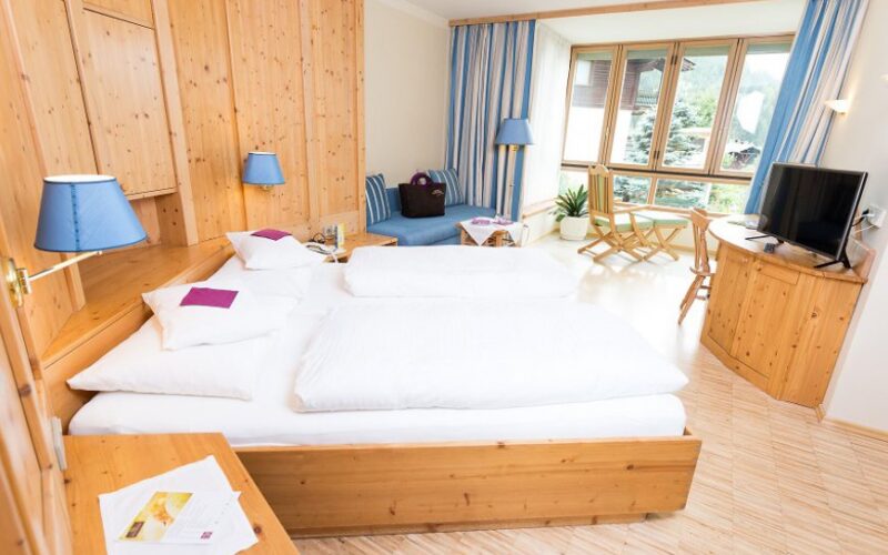 Ein Hotelzimmer mit einem Doppelbett, Holzmöbeln, einem Fernseher, einer kleinen Sitzecke an großen Fenstern und blauen Akzenten in Lampen und Vorhängen.