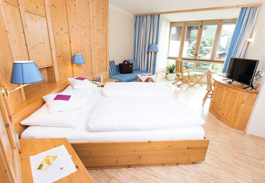 Ein Hotelzimmer mit einem Doppelbett, Holzmöbeln, einem Fernseher, einer kleinen Sitzecke an großen Fenstern und blauen Akzenten in Lampen und Vorhängen.