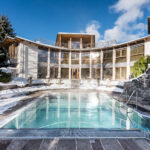 Freibad, umgeben von einem geschwungenen, modernen Gebäude, mit schneebedeckten Bäumen und Boden, unter einem klaren blauen Himmel.