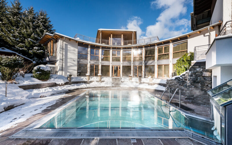 Freibad, umgeben von einem geschwungenen, modernen Gebäude, mit schneebedeckten Bäumen und Boden, unter einem klaren blauen Himmel.
