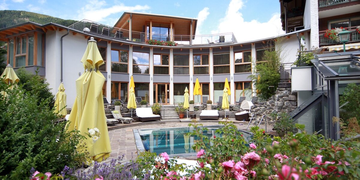 Ein geschwungenes, modernes Hotelgebäude mit großen Fenstern blickt auf einen kreisrunden Außenpool, der von gelben Sonnenschirmen, Liegestühlen und bunt blühenden Pflanzen umgeben ist.