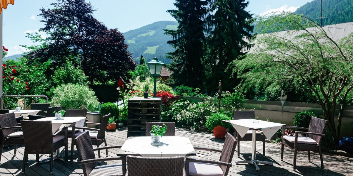 Terrasse mit Korbstühlen und Tischen auf einer Holzterrasse, umgeben von üppigem Grün und der Berglandschaft im Hintergrund.