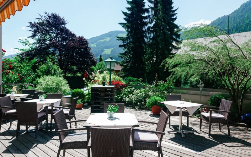 Terrasse mit Korbstühlen und Tischen auf einer Holzterrasse, umgeben von üppigem Grün und der Berglandschaft im Hintergrund.