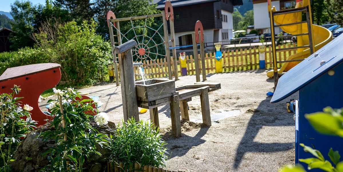 Spielplatz im Freien mit hölzernen Klettergerüsten, Rutsche, Bänken und Spielgeräten auf Sand, umgeben von Grünflächen und Häusern im Hintergrund.