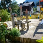Spielplatz im Freien mit hölzernen Klettergerüsten, Rutsche, Bänken und Spielgeräten auf Sand, umgeben von Grünflächen und Häusern im Hintergrund.