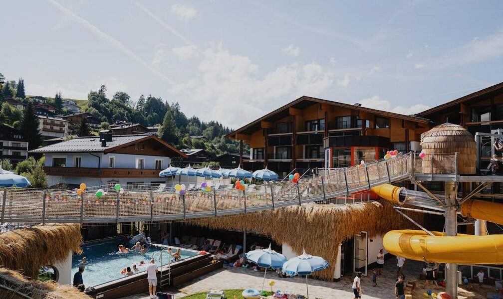 Außenpoolbereich eines Resorts mit Menschen, die schwimmen, auf Sonnenliegen liegen und Kinder, die die gelben Wasserrutschen benutzen. Im Hintergrund sind Sonnenschirme, Luftballons und Gebäude im Chalet-Stil zu sehen.