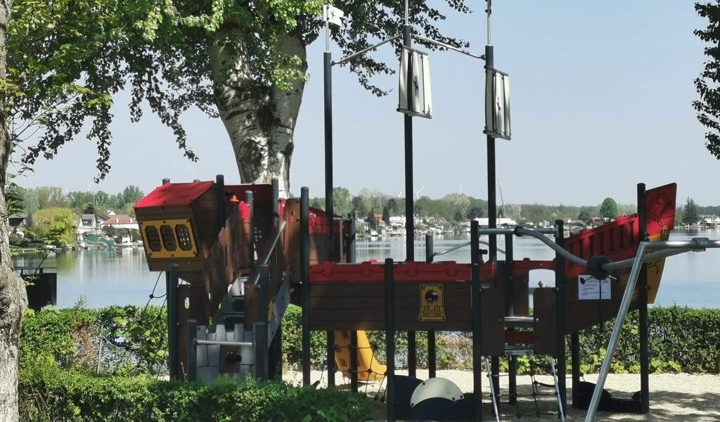 Ein Spielplatz in Form eines Piratenschiffs befindet sich in der Nähe eines Sees, umgeben von Bäumen und Grünflächen, mit Häusern auf der anderen Seite des Wassers.