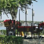 Ein Spielplatz in Form eines Piratenschiffs befindet sich in der Nähe eines Sees, umgeben von Bäumen und Grünflächen, mit Häusern auf der anderen Seite des Wassers.