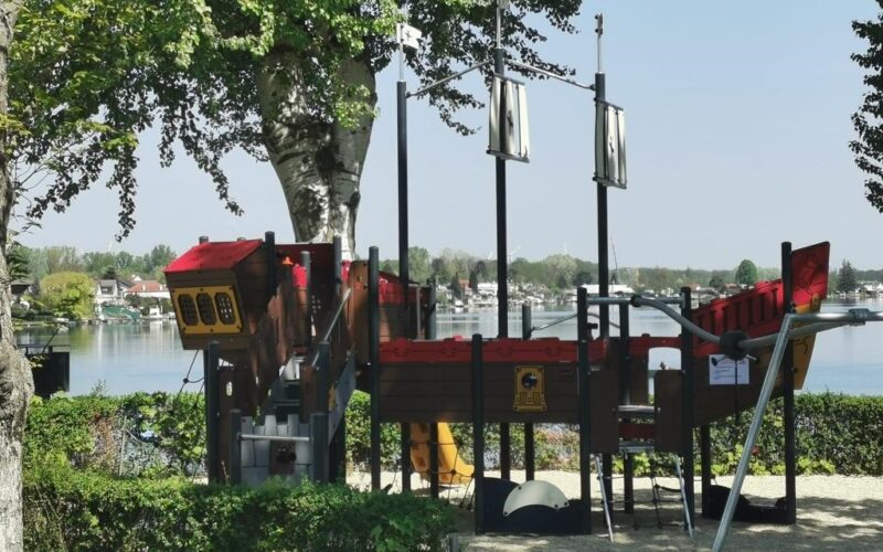 Ein Spielplatz in Form eines Piratenschiffs befindet sich in der Nähe eines Sees, umgeben von Bäumen und Grünflächen, mit Häusern auf der anderen Seite des Wassers.