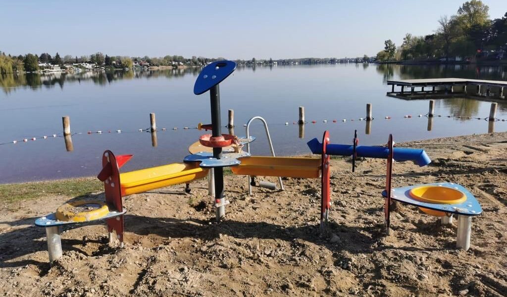 Ein Sand- und Wasserspielplatz für Kinder an einem sandigen Seeufer mit ruhigem Wasser, einem Steg und Häusern im Hintergrund.