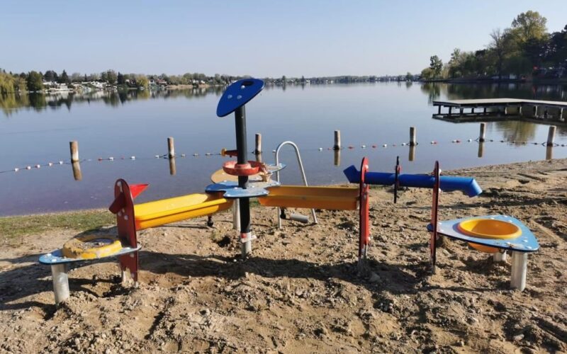 Ein Sand- und Wasserspielplatz für Kinder an einem sandigen Seeufer mit ruhigem Wasser, einem Steg und Häusern im Hintergrund.