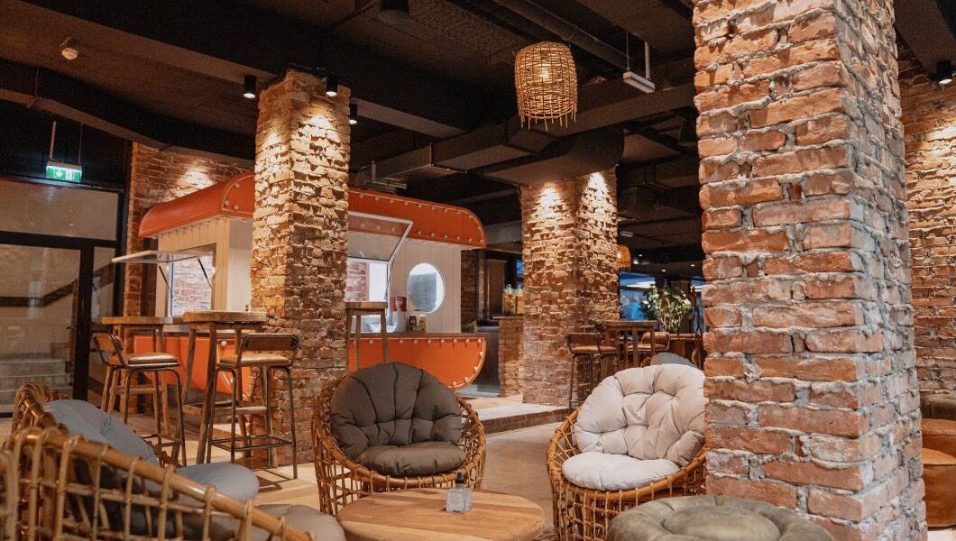 Eine moderne Lounge mit freiliegenden Backsteinsäulen, Korbstühlen, runden Tischen und einer orangefarbenen Foodtruck-Theke im Hintergrund.
