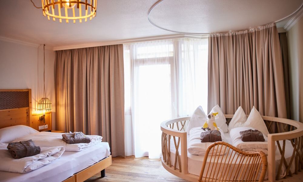 Ein Hotelzimmer mit einem Doppelbett, einem runden Bett im Stil eines Kinderbettes, Holzfußboden, einem Korbstuhl und großen Fenstern mit beigen Vorhängen.