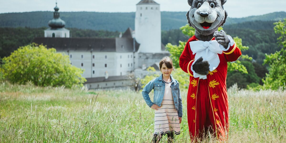 Ein junges Mädchen steht neben einer Person in einem schwarzen Tierkostüm, die einen roten Mantel trägt, mit einer Burg und Bäumen im Hintergrund.
