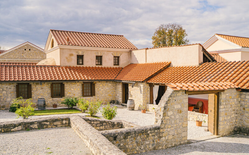 Steinhof mit Topfpflanzen und Fässern, umgeben von Gebäuden mit roten Ziegeldächern unter einem teilweise bewölkten Himmel.