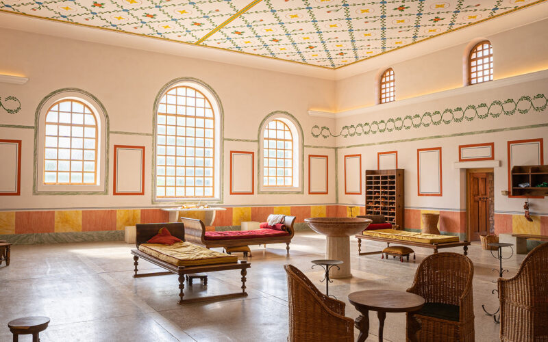 Ein geräumiger Raum mit gewölbten Fenstern, dekorativer Decke und Wänden, Liegestühlen, Korbstühlen, einem zentralen Steinbecken und Holzregalen an einer Wand.