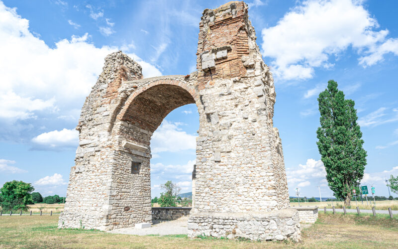 Die Ruinen eines antiken römischen Bogens aus Stein und Ziegeln stehen auf einem grasbewachsenen Feld unter blauem Himmel, umgeben von Bäumen und fernen Zeichen.