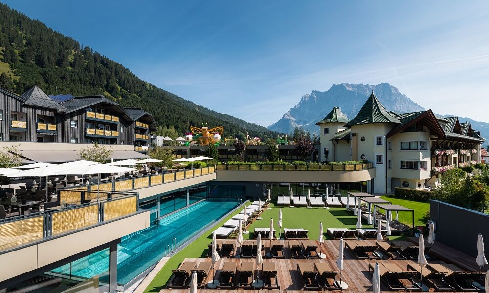 Luxushotel mit Außenpool, Liegestühlen und Terrasse inmitten von bewaldeten Hügeln und einem Berg unter blauem Himmel.