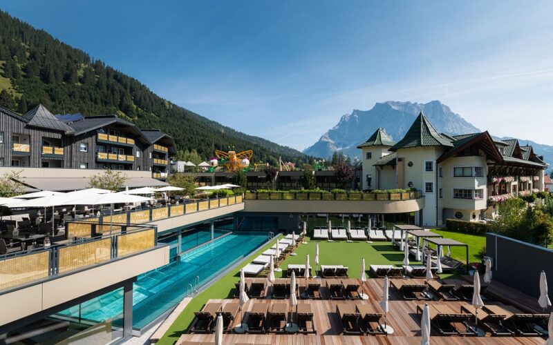 Luxushotel mit Außenpool, Liegestühlen und Terrasse inmitten von bewaldeten Hügeln und einem Berg unter blauem Himmel.