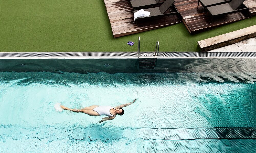 Eine Person in einem weißen Badeanzug schwimmt auf dem Rücken in einem Schwimmbad, während auf einem Deck in der Nähe leere Liegestühle und Handtücher zu sehen sind.