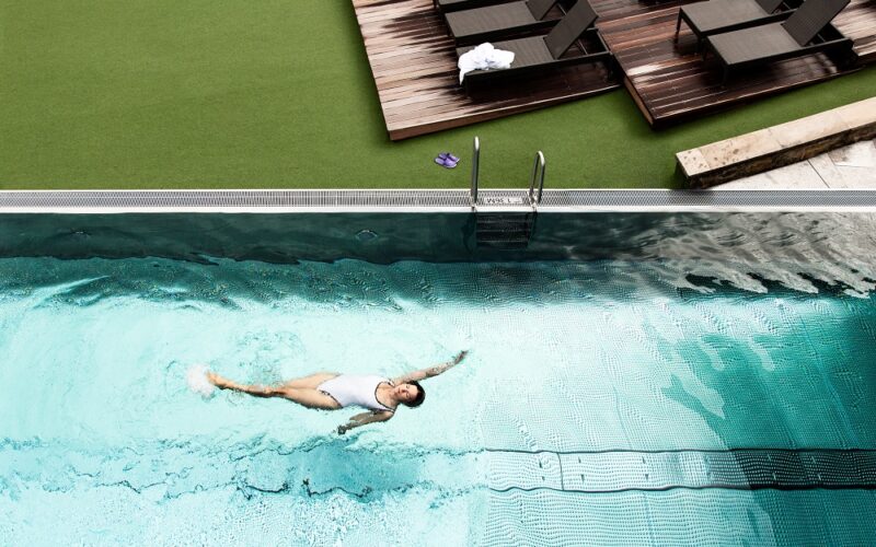 Eine Person in einem weißen Badeanzug schwimmt auf dem Rücken in einem Schwimmbad, während auf einem Deck in der Nähe leere Liegestühle und Handtücher zu sehen sind.