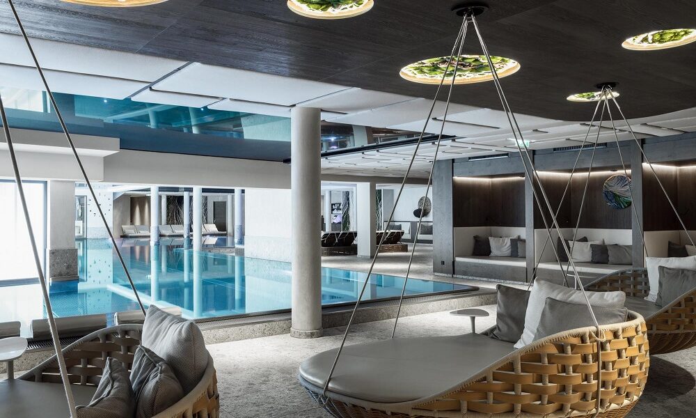 Moderner Hallenbadbereich mit großen gepolsterten Schaukelstühlen, Glaswänden, einem Becken mit blauem Wasser und Deckenleuchten, die einen eleganten Freizeitbereich schaffen.