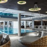 Moderner Hallenbadbereich mit großen gepolsterten Schaukelstühlen, Glaswänden, einem Becken mit blauem Wasser und Deckenleuchten, die einen eleganten Freizeitbereich schaffen.