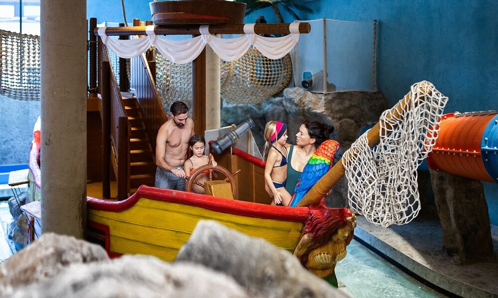 Vier Personen, darunter zwei Kinder und zwei Erwachsene in Badeanzügen, stehen auf einem Indoor-Spielplatz, der die Form eines Piratenschiffs hat, mit Netzen und Rutschen um sie herum.