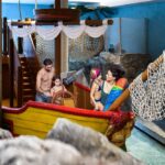 Vier Personen, darunter zwei Kinder und zwei Erwachsene in Badeanzügen, stehen auf einem Indoor-Spielplatz, der die Form eines Piratenschiffs hat, mit Netzen und Rutschen um sie herum.