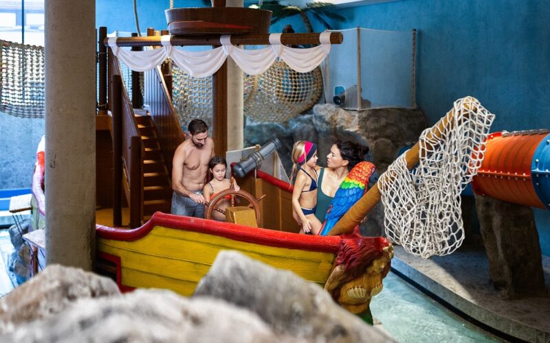 Vier Personen, darunter zwei Kinder und zwei Erwachsene in Badeanzügen, stehen auf einem Indoor-Spielplatz, der die Form eines Piratenschiffs hat, mit Netzen und Rutschen um sie herum.