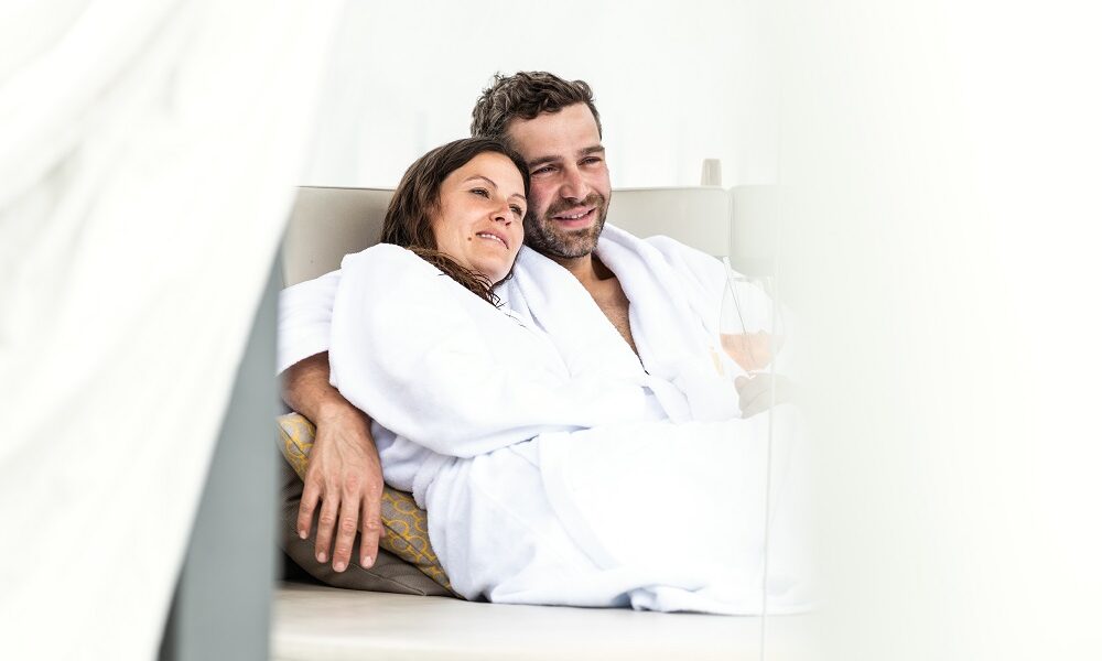 Ein Paar in weißen Bademänteln sitzt eng beieinander auf einer Couch, lächelt und entspannt sich.