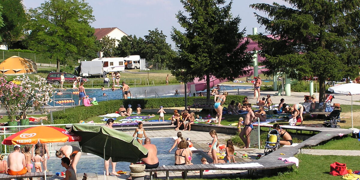 Menschen verschiedenen Alters schwimmen, sitzen und entspannen sich an einem sonnigen Tag um ein Freibad in einer Parkanlage mit Bäumen und Sonnenschirmen.