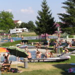Menschen verschiedenen Alters schwimmen, sitzen und entspannen sich an einem sonnigen Tag um ein Freibad in einer Parkanlage mit Bäumen und Sonnenschirmen.