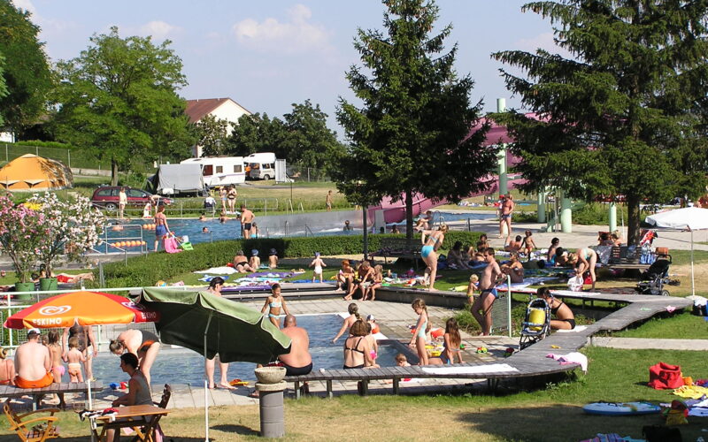 Menschen verschiedenen Alters schwimmen, sitzen und entspannen sich an einem sonnigen Tag um ein Freibad in einer Parkanlage mit Bäumen und Sonnenschirmen.