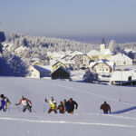 Eine Gruppe von Langläufern auf einem verschneiten Feld in der Nähe eines Dorfes mit schneebedeckten Häusern und einer Kirche im Hintergrund.