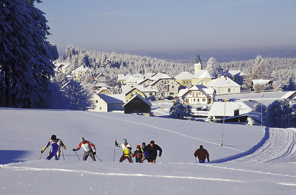 Eine Gruppe von Langläufern auf einem verschneiten Feld in der Nähe eines Dorfes mit schneebedeckten Häusern und einer Kirche im Hintergrund.