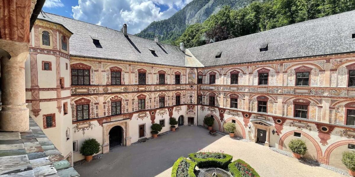 Ein Innenhof mit einem runden Gartenmittelstück, umgeben von einem dreistöckigen historischen Gebäude mit verzierten Fassaden und Bogenfenstern, vor der Kulisse von Bergen und einem teilweise bewölkten Himmel.