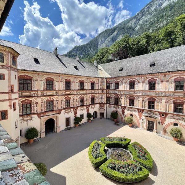 Ein Innenhof mit einem runden Gartenmittelstück, umgeben von einem dreistöckigen historischen Gebäude mit verzierten Fassaden und Bogenfenstern, vor der Kulisse von Bergen und einem teilweise bewölkten Himmel.