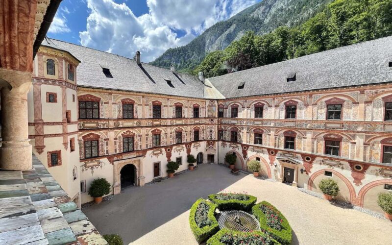 Ein Innenhof mit einem runden Gartenmittelstück, umgeben von einem dreistöckigen historischen Gebäude mit verzierten Fassaden und Bogenfenstern, vor der Kulisse von Bergen und einem teilweise bewölkten Himmel.