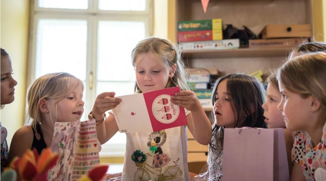 Eine Gruppe kleiner Kinder sitzt um einen Tisch herum, während ein Mädchen in der Mitte eine Grußkarte öffnet und vorliest.