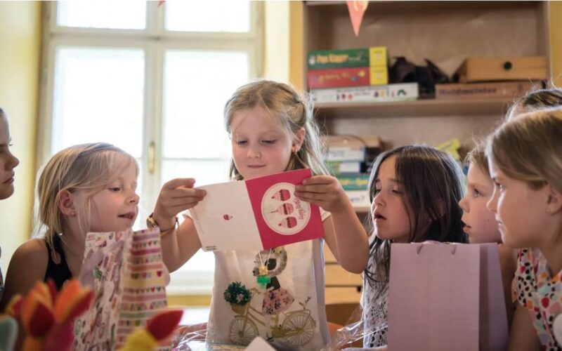 Eine Gruppe kleiner Kinder sitzt um einen Tisch herum, während ein Mädchen in der Mitte eine Grußkarte öffnet und vorliest.