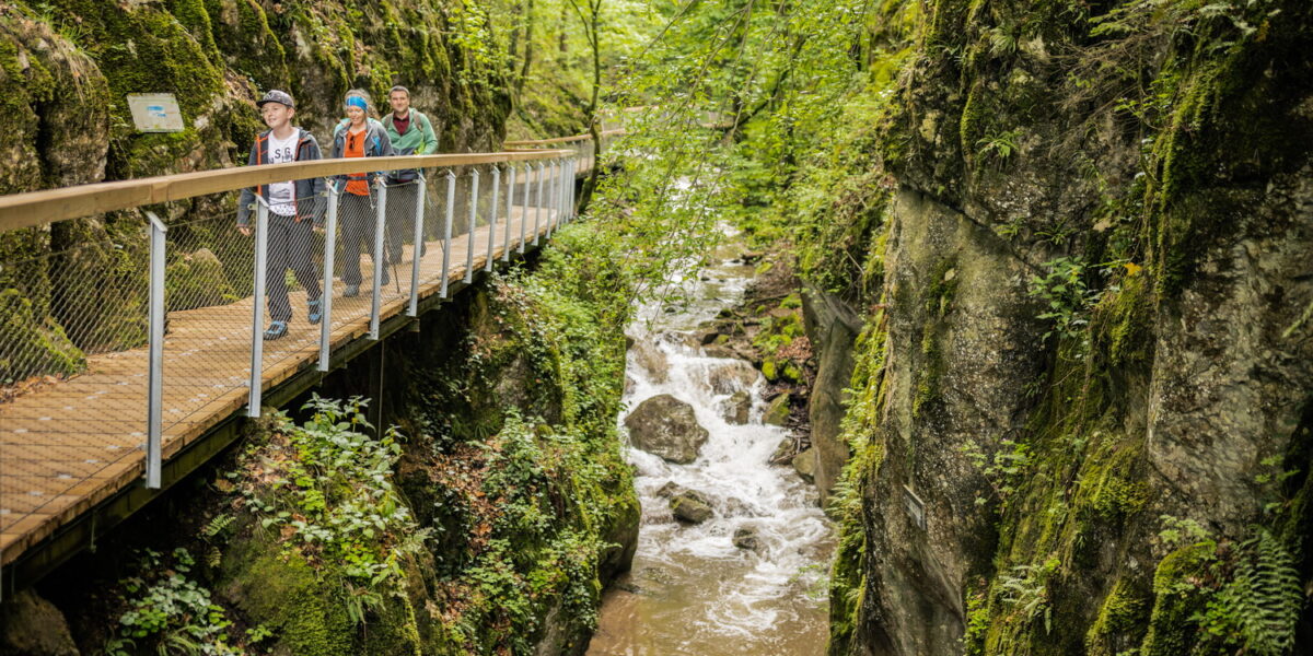 Drei Personen gehen auf einer Holzbrücke über eine enge, felsige Schlucht mit einem fließenden Bach, umgeben von dichter grüner Vegetation.