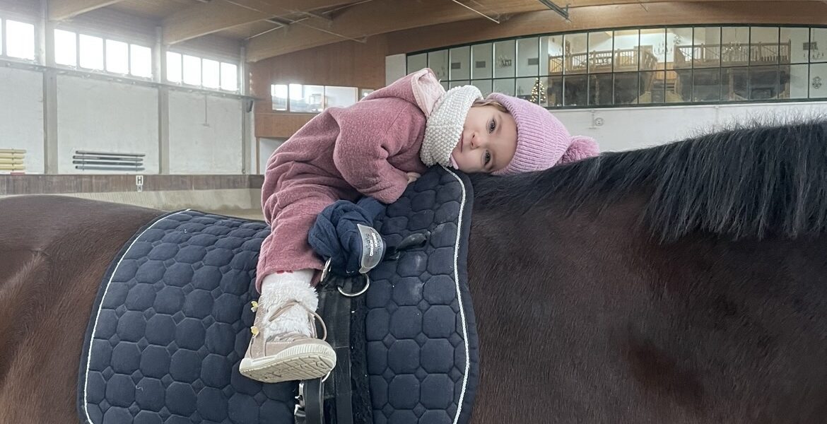 Ein kleines Kind, warm angezogen und mit einer rosa Mütze, liegt auf dem Bauch auf einem gesattelten braunen Pferd in einer Reithalle.