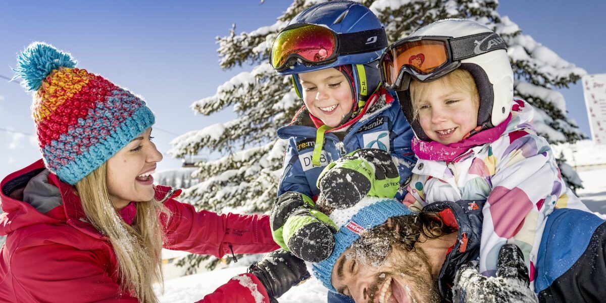 Eine Frau und drei Kinder in Winterkleidung lachen und spielen zusammen im Schnee mit einem verschneiten Baum im Hintergrund.