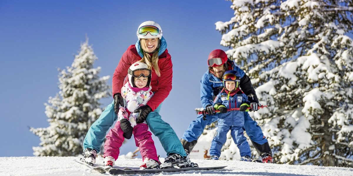 Eine vierköpfige Familie in Winterkleidung fährt gemeinsam auf einer verschneiten Piste mit schneebedeckten Bäumen und einem klaren blauen Himmel im Hintergrund Ski.