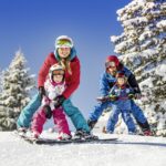 Eine vierköpfige Familie in Winterkleidung fährt gemeinsam auf einer verschneiten Piste mit schneebedeckten Bäumen und einem klaren blauen Himmel im Hintergrund Ski.