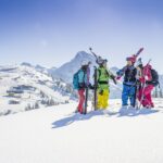Vier Personen in farbenfroher Skiausrüstung stehen mit Skiern und Stöcken auf einem verschneiten Berghang, mit schneebedeckten Gipfeln und einem klaren blauen Himmel im Hintergrund.