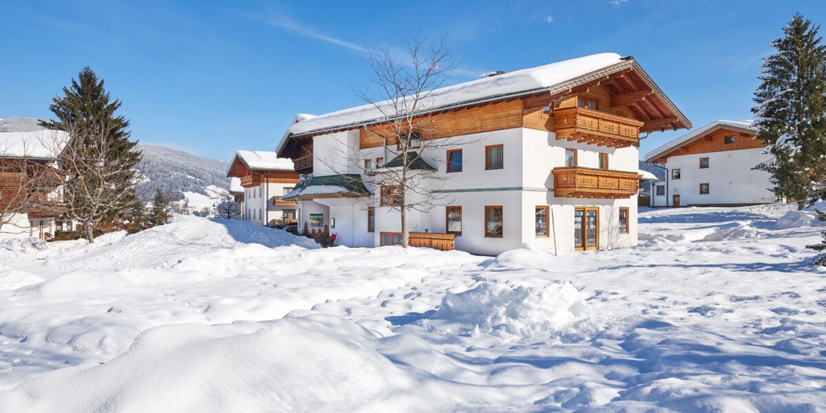 Ein Haus im Chalet-Stil mit Holzbalkonen liegt inmitten von tiefem Schnee unter einem klaren blauen Himmel, mit Bergen und Bäumen im Hintergrund.