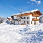 Ein Haus im Chalet-Stil mit Holzbalkonen liegt inmitten von tiefem Schnee unter einem klaren blauen Himmel, mit Bergen und Bäumen im Hintergrund.